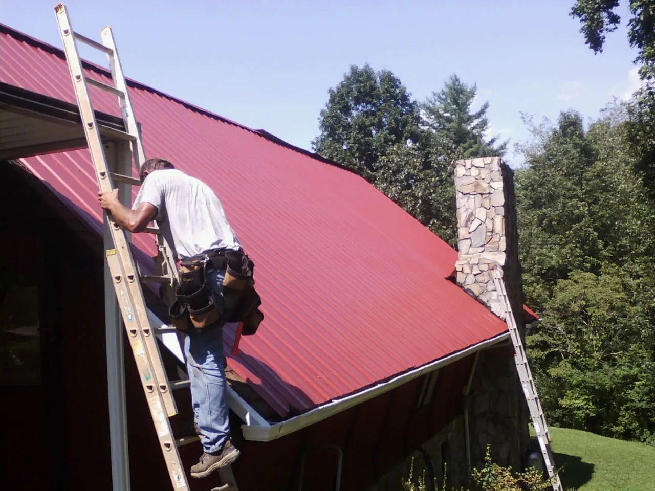 Roofer on a ladder beside a red metal roof and stone chimney — Soffit & Fascia Repair by Roofing Brothers in Rosamond